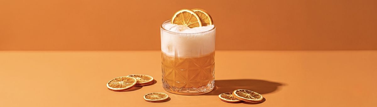 Minimalistisches Food-Foto im Panorama-Format von einem spritzigen orangefarbenen Kombucha-Drink im gemusterten Glas mit weißer Schaumkrone, garniert mit getrockneten Orangenscheiben auf einem nahtlosen, orangefarbenen Studio-Hintergrund