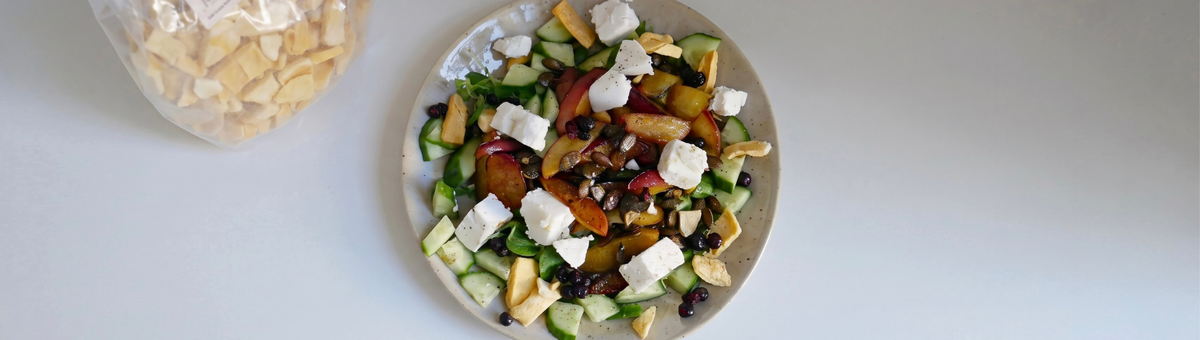 Draufsicht auf einen Salat mit gegrillten Pfirsichen, veganem Feta, Gurken und Kürbiskernen, bestreut mit gefriergetrockneten Mangostücken; oben links steht eine transparente Tüte mit KoRo Mangostücken.