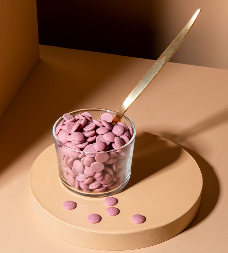 Rosafarbene Schoko-Drops aus Ruby-Kakaobohnen in einem kleinen Glas mit einem goldenen Löffel auf hellem Untergrund