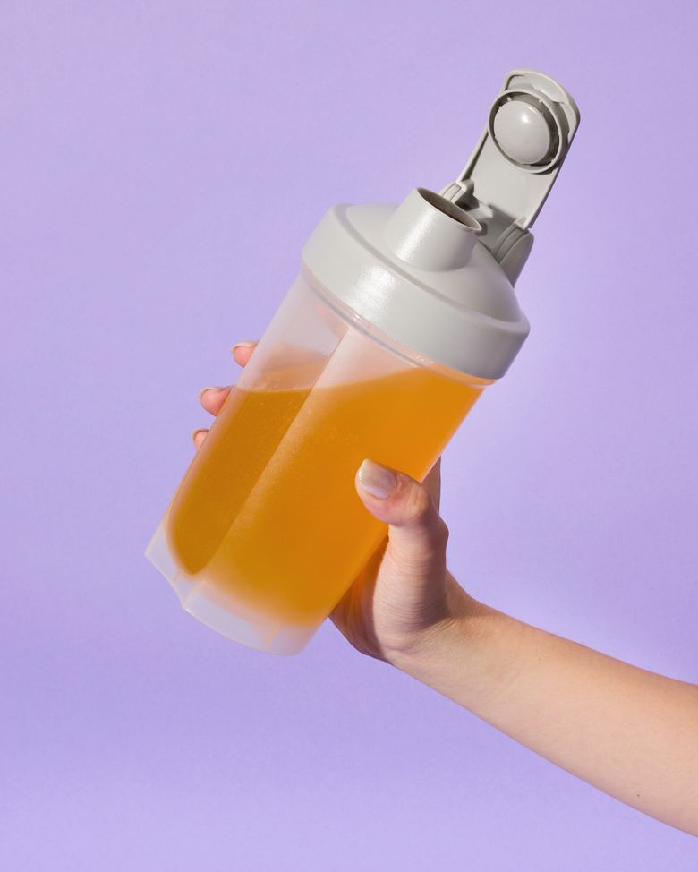 Hand hält einen mit Clear Protein halb gefüllten, durchsichtigen Proteinshaker mit grauem Deckel vor einem lilafarbenen Hintergrund.