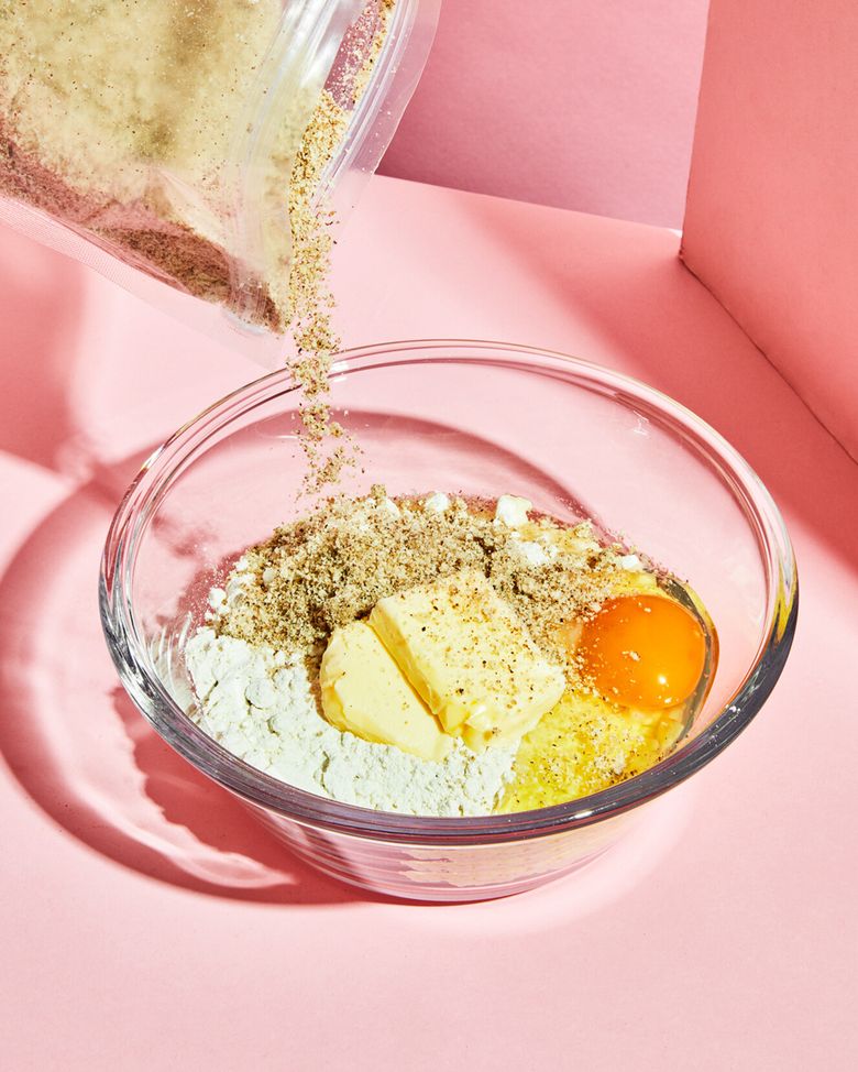 Eine Glas-Rührschüssel auf pinkem Hintergrund, gefüllt mit Backzutaten: Mehl, einem Ei, zwei Stücke Butter und eine helle Pulvermischung. Von oben wird weiteres bräunliches Pulver, vermutlich Proteinpulver, aus einem durchsichtigen Beutel in die Schüssel geschüttet.