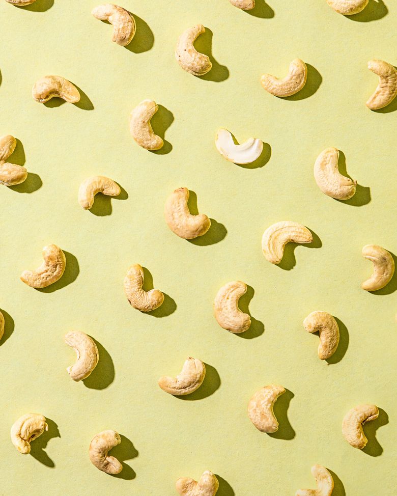 Einzelne Cashewkerne liegen in Reihen angeordnet auf einem hellgrünen Hintergrund. Die Cashewkerne sind ungeröstet und haben eine helle, cremeweiße Farbe. Sie werfen durch eine starke Lichtquelle von oben rechts Schatten.