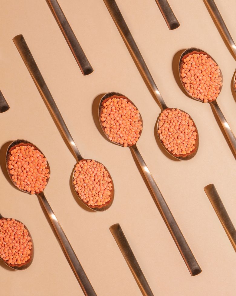 Mehrere glänzende, metallische Löffel, die diagonal von unten links nach oben rechts über eine beige Oberfläche angeordnet sind. Jeder Löffel ist gefüllt mit kleinen, roten Linsen.