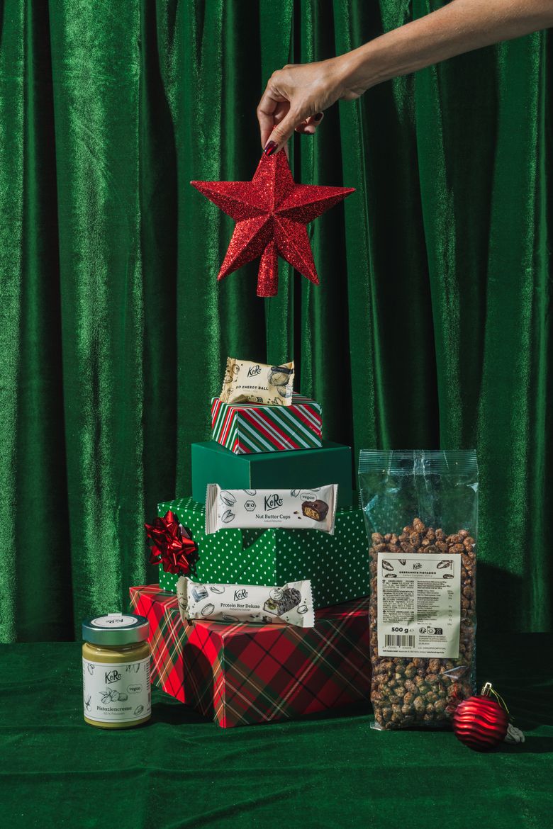 Eine festliche Weihnachtsinszenierung vor grünem Samthintergrund. Geschenkboxen in verschiedenen Grüntönen und Rot, gestapelt in Form eines Tannenbaums mit einem roten Stern als Spitze, umgeben von veganen Koro-Produkten wie Nussmus, Proteinriegeln, Nut Butter Cups und gerösteten Kichererbsen.