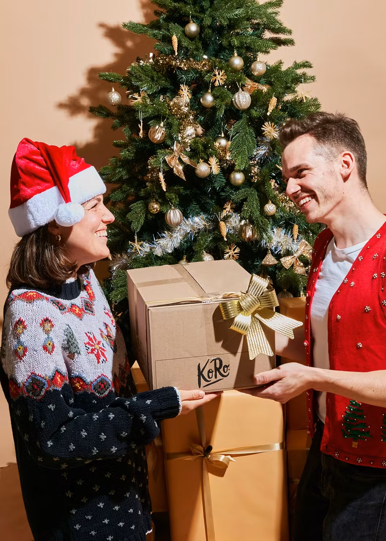 Ein lachendes Paar in weihnachtlicher Kleidung, das gemeinsam ein großes, in braunem Karton verpacktes Geschenk mit dem "KoRo"-Logo und einer goldenen Schleife hält. Die Frau trägt eine Nikolausmütze und einen Norwegerpullover, der Mann einen roten Strick-Pullunder mit Weihnachtsmotiven. Hinter ihnen steht ein geschmückter Weihnachtsbaum und eine Stapel weiterer, in Goldpapier verpackter Geschenke.