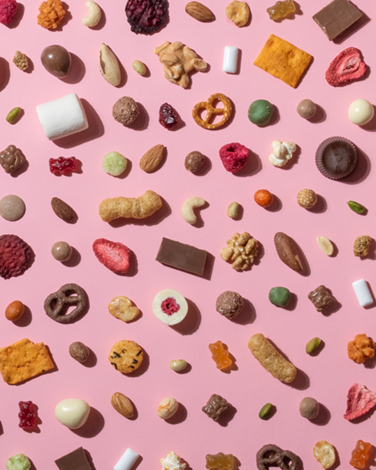  Eine vielfältige Auswahl an Snacks, darunter Nüsse, Gummibärchen, Schokolade, Brezeln und getrocknete Früchte, ist in einem gleichmäßigen Raster auf einem hellrosa Hintergrund angeordnet. 