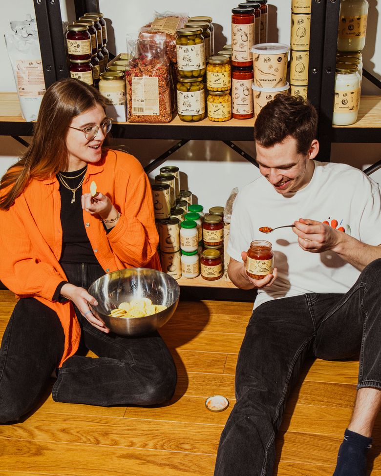 Zwei Personen sitzen entspannt vor KoRo-Produktregalen und probieren fröhlich verschiedene Snacks und Aufstriche.