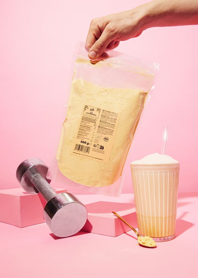 Eine Hand hält eine Packung KoRo Bio Lupinenmehl neben einer Hantel und einem Protein-Shake auf rosa Hintergrund.