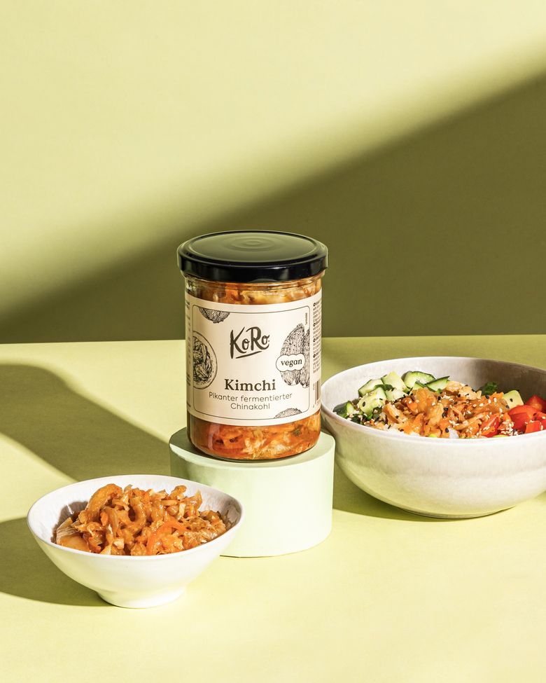 Glas mit Kimchi als Beispiel für fermentierte Lebensmittel, daneben eine Schale mit fermentiertem Kohl und eine Bowl mit frischem Gemüse auf hellgrünem Hintergrund.