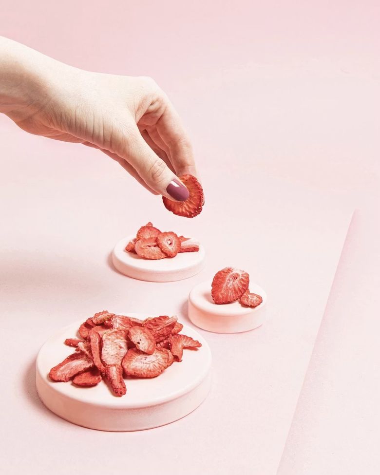 Eine Hand greift nach einer gefriergetrockneten Erdbeerscheibe. Darunter liegen weitere getrocknete Erdbeerscheiben als Snack arrangiert auf drei kleinen, rosa Podesten auf einem rosa Hintergrund.