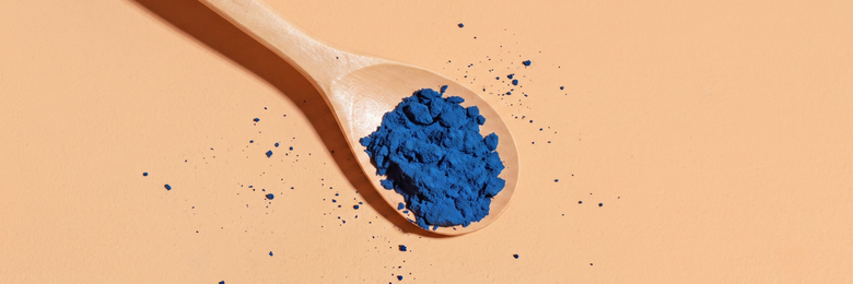 Ein Holzlöffel voller blaues Spirulina-Pulver auf einem hell-orangen Hintergrund. Nahaufnahme von oben.