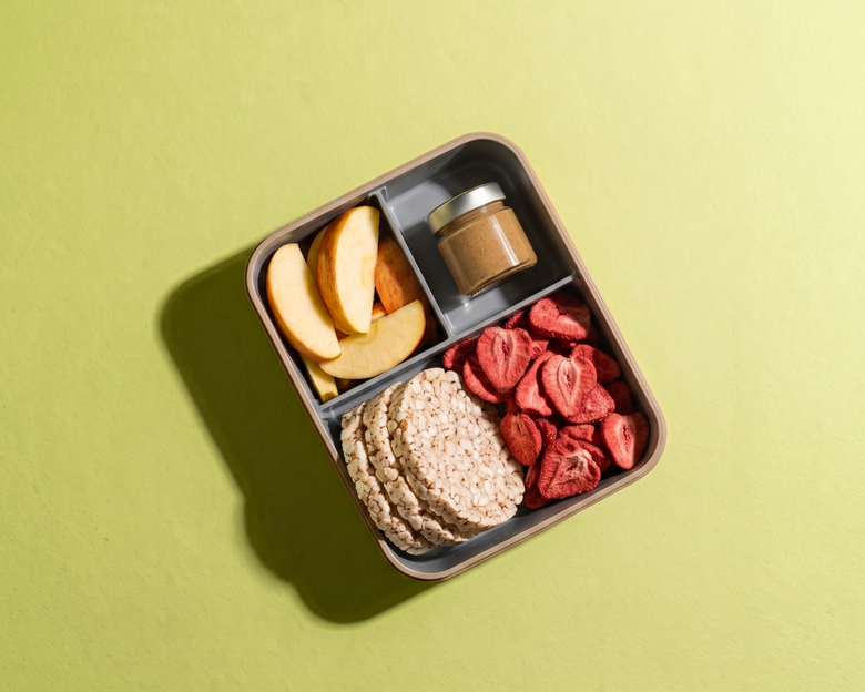 Draufsicht auf eine strukturierte Bento-Box im Querformat auf hellgrünem Hintergrund, befüllt mit frischen Apfelscheiben, einem kleinen Glas Nussmus, runden Reiswaffeln und gefriergetrockneten Erdbeerscheiben als Snack.