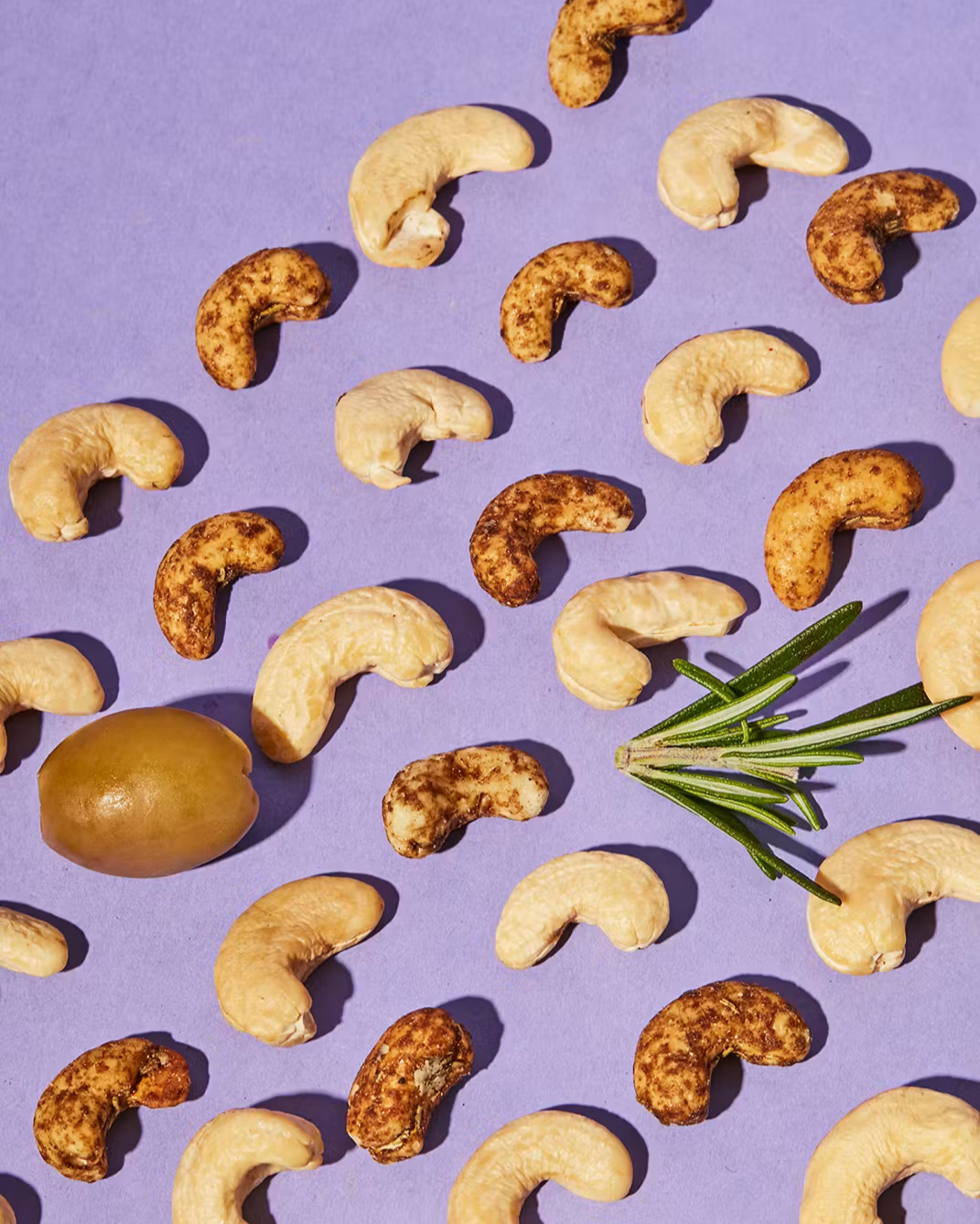 Draufsicht auf verstreute Cashewkerne – teils naturbelassen hell, teils mit einer braunen Gewürzmischung überzogen – auf einem lila Hintergrund, ergänzt durch eine grüne Olive und einen Rosmarinzweig.