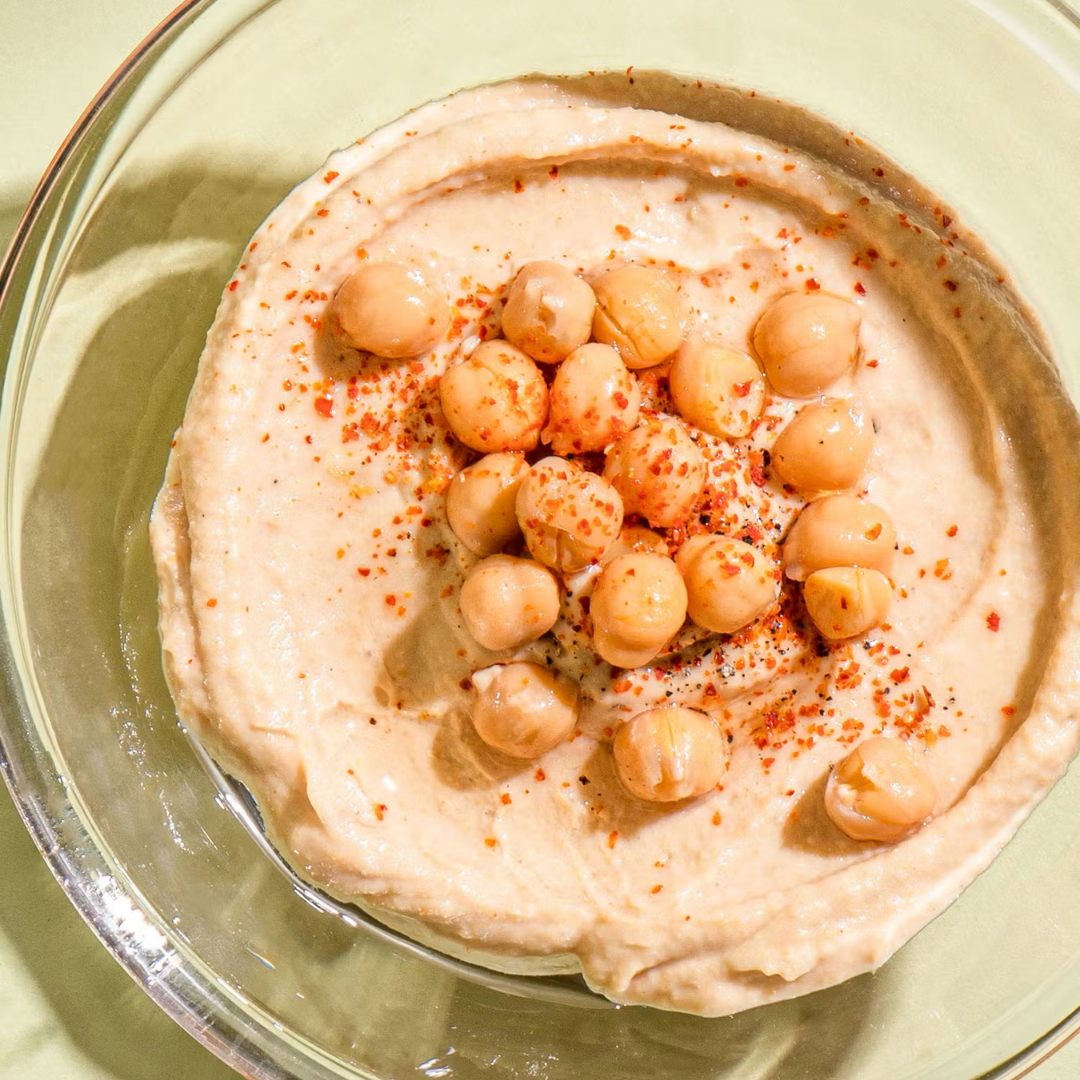 Gemini hat gesagt Eine Glasschale mit cremigem Hummus, garniert mit ganzen Kichererbsen und roten Gewürzen.