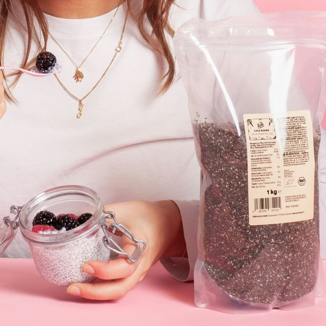 Eine Person hält ein Glas Chia-Pudding mit frischen Beeren neben einer 1-kg-Packung KoRo Chia-Samen auf rosa Hintergrund.