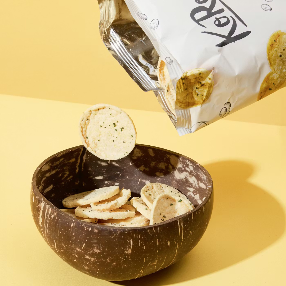 Linsenchips werden aus einer KoRo-Packung in eine dunkle Kokosnussschale auf gelbem Hintergrund geschüttet.