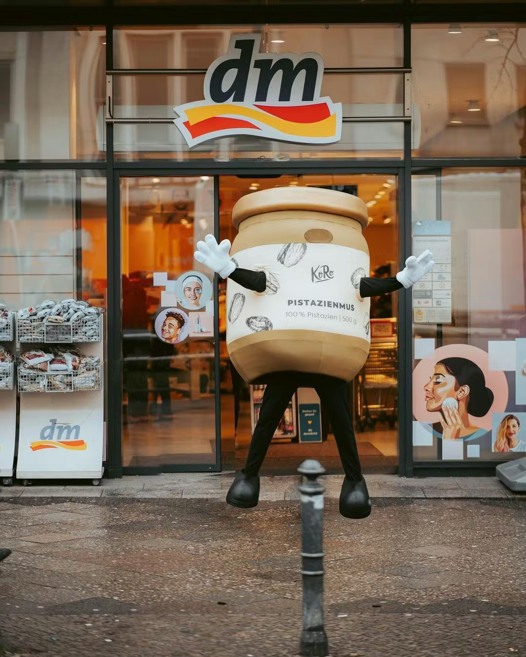 Eine Person in einem lebensgroßen KoRo-Pistazienmus-Glas-Kostüm springt mit ausgebreiteten Armen vor dem Eingang einer dm-Drogeriemarkt-Filiale in die Luft.