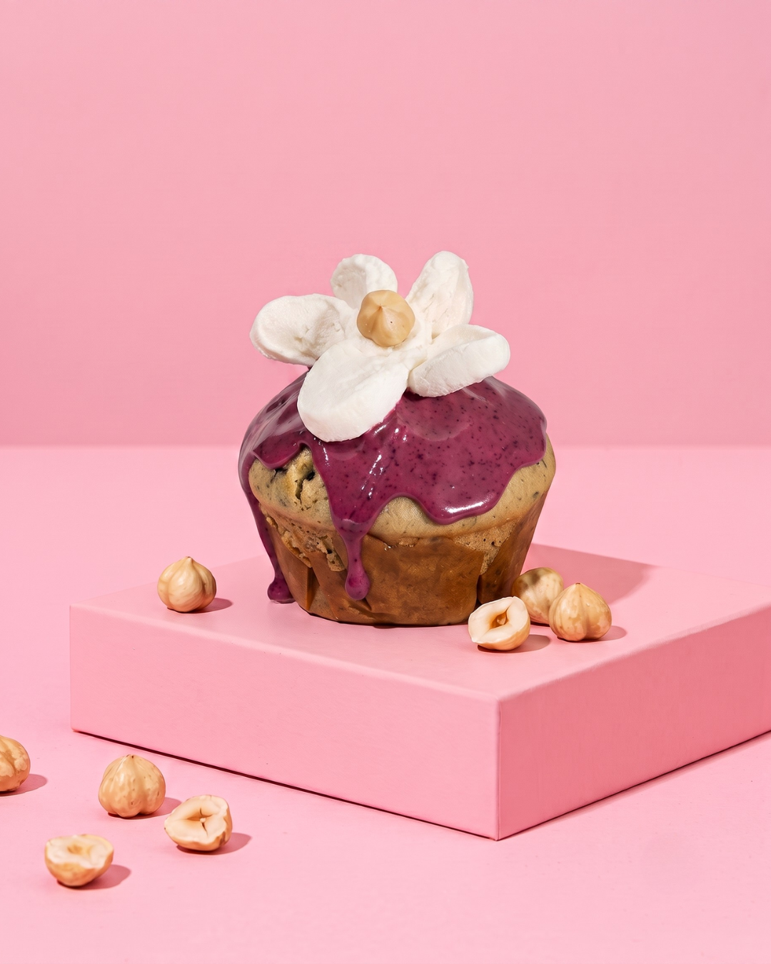 Saftiger Blaubeer-Muffin mit lilafarbenem Heidelbeer-Guss, verziert mit einer weißen Marshmallow-Blüte und einer Haselnuss auf einem rosa Podest vor pinkem Hintergrund.