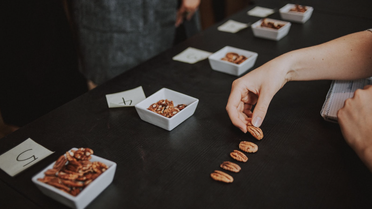 Sur une table noire se trouvent plusieurs petits bols de noix du Brésil, numérotés par des bouts de papier devant eux. Une main dispose quelques noix les unes à côté des autres.