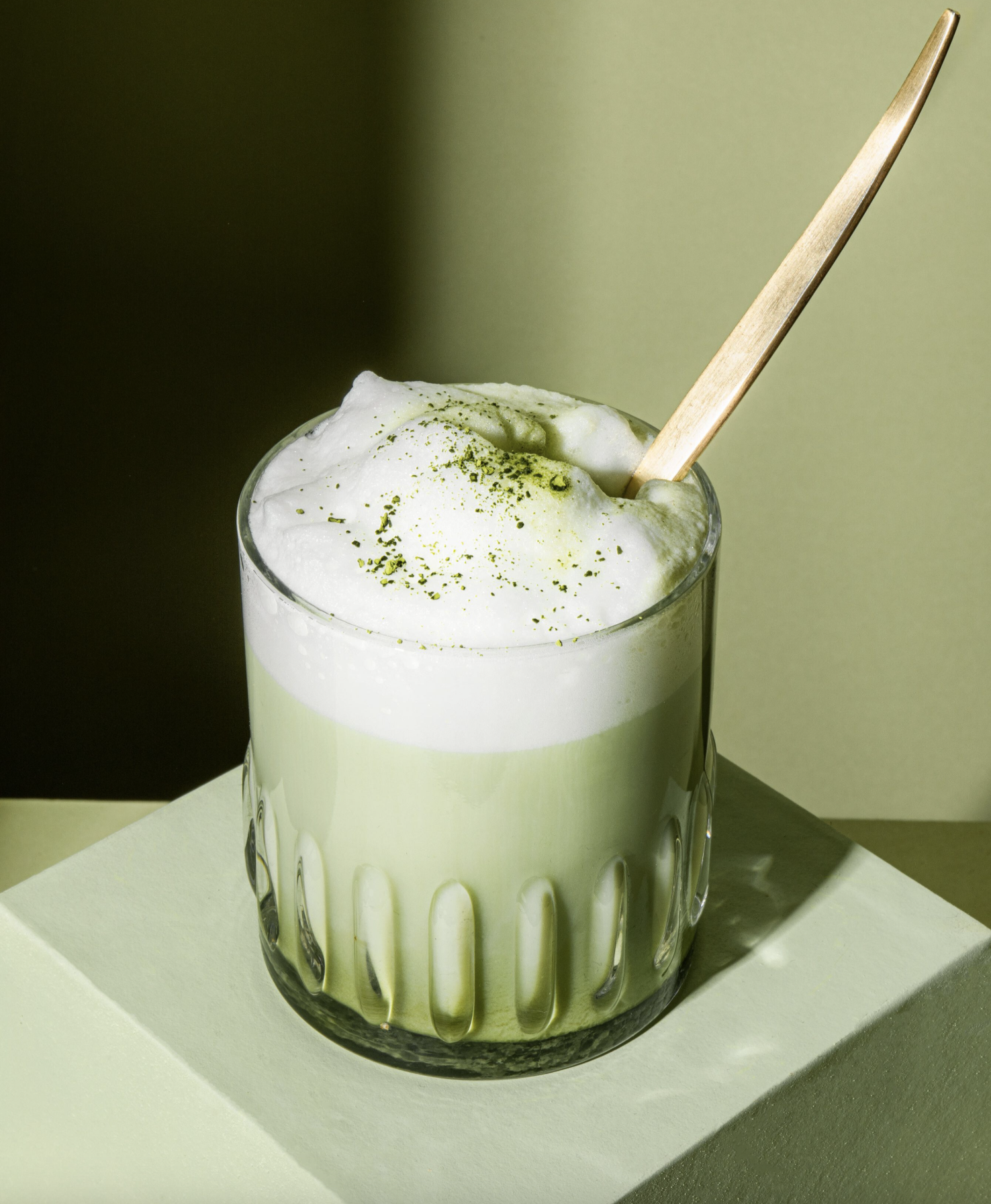 Un verre transparent et strié contenant un latte matcha vert surmonté d'une couche de mousse blanche épaisse et de quelques grains de poudre de matcha.