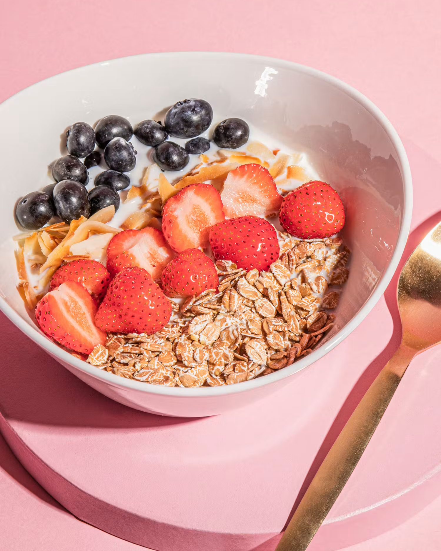 Nahaufnahme: Weiße Schale gefüllt mit Bio-Dinkelflocken Großblatt, Pflanzendrink, frischen Erdbeeren, Heidelbeeren und Kokoschips auf rosafarbenem Untergrund.