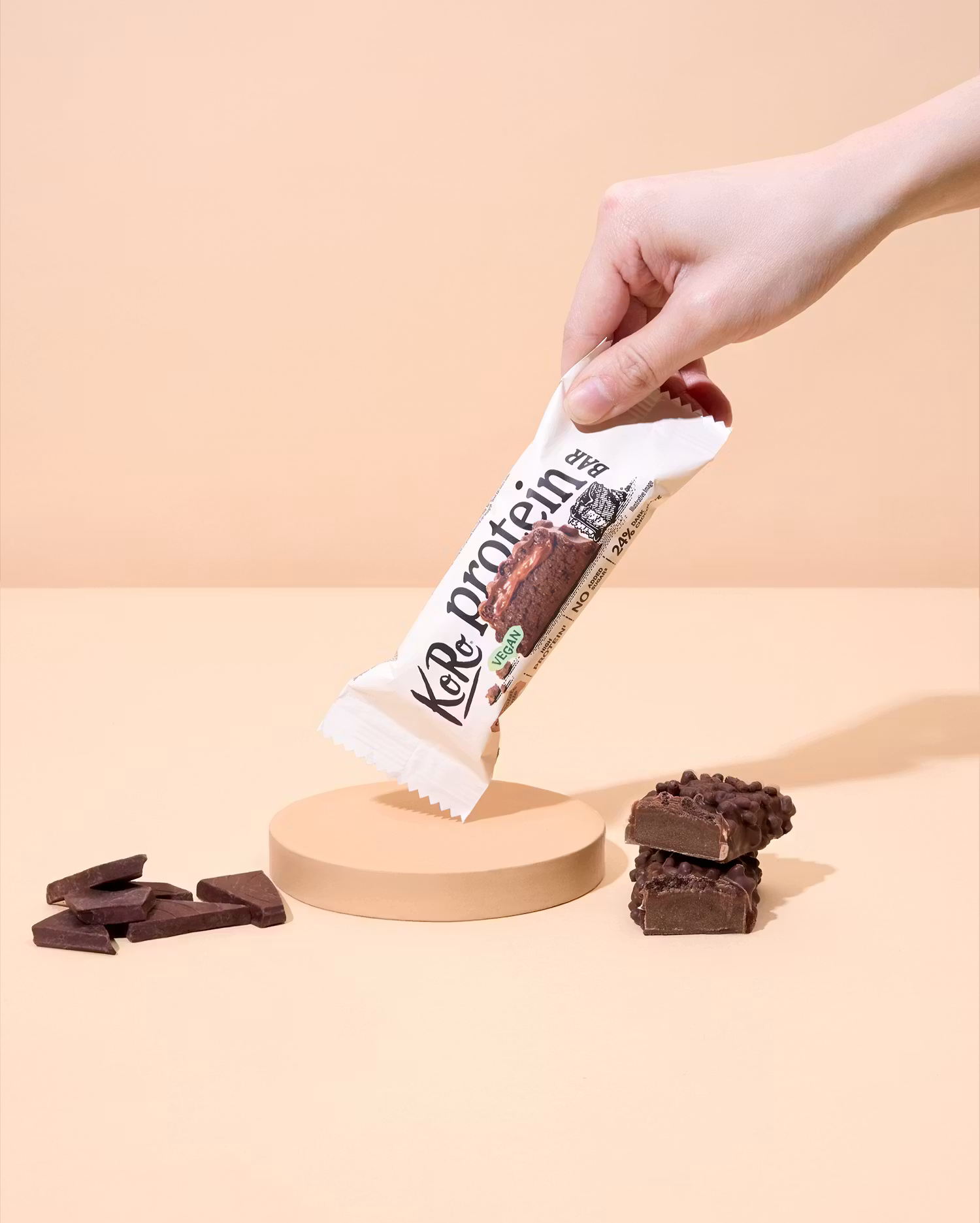 Eine Hand hält einen verpackten KoRo Protein Bar über einem Podest, umgeben von Schokoladenstücken auf hellem Untergrund.