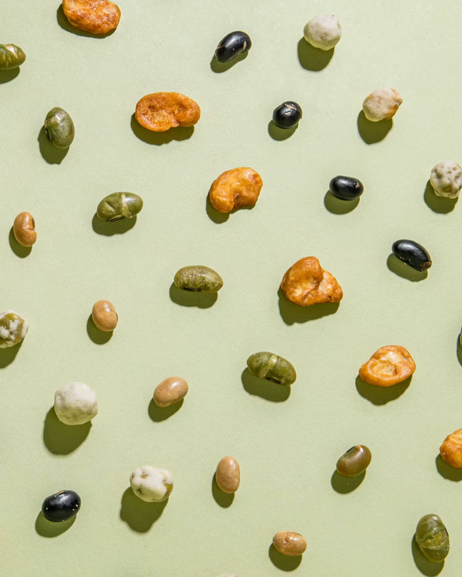 Eine bunte Auswahl an verschiedenen gerösteten Bohnen und Knabber-Snacks, die ordentlich und mit klaren Schatten auf einem hellgrünen Hintergrund verteilt sind.