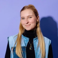 Flavie lächelt in die Kamera, sie trägt ein schwarzes Oberteil und eine Jeansweste sowie große Ohrringe und eine Kette