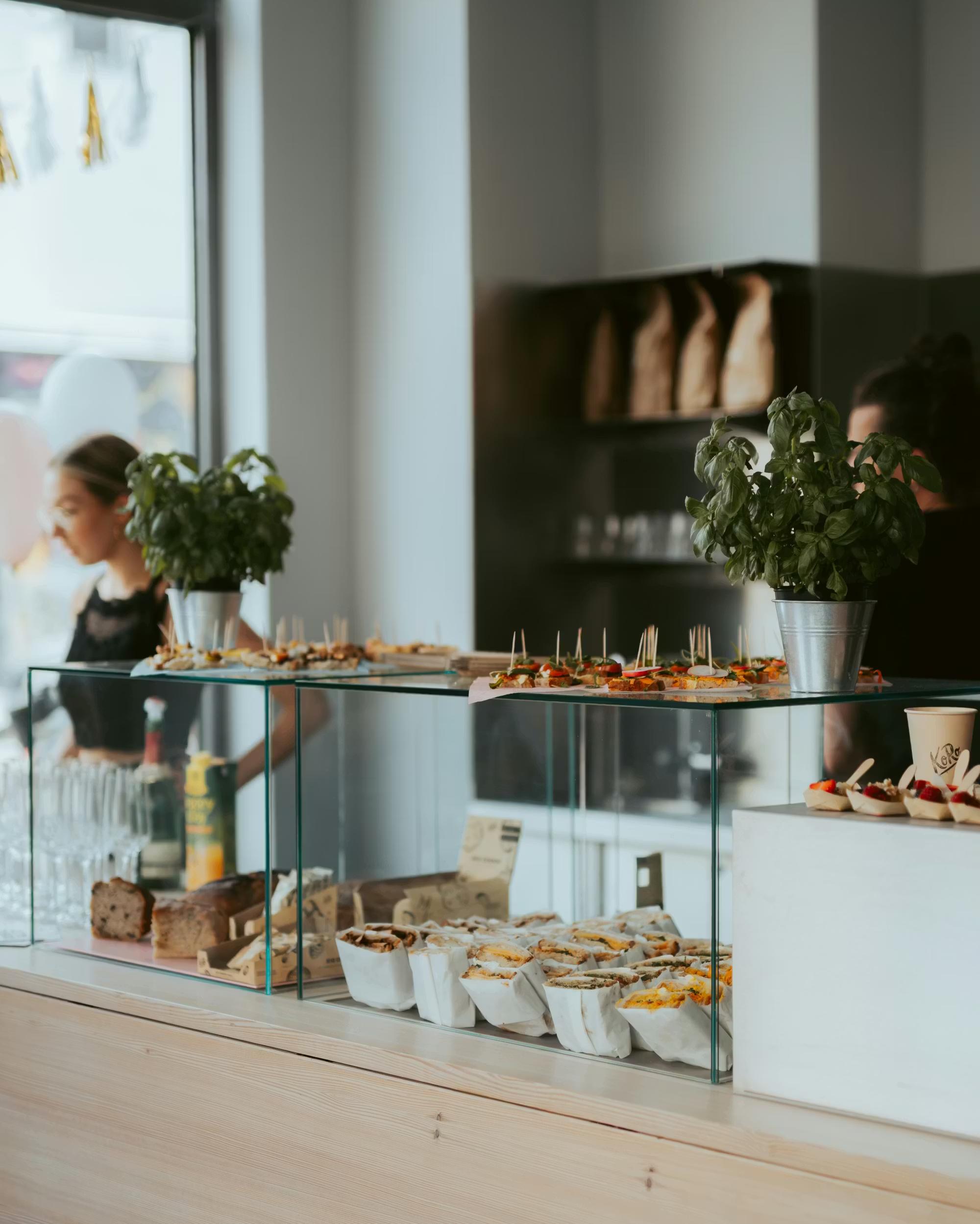 Blick auf die moderne Verkaufstheke im KoRo Café mit einer Auswahl an belegten Stullen, frischem Gebäck und Basilikumpflanzen in Zinktöpfen als Dekoration.
