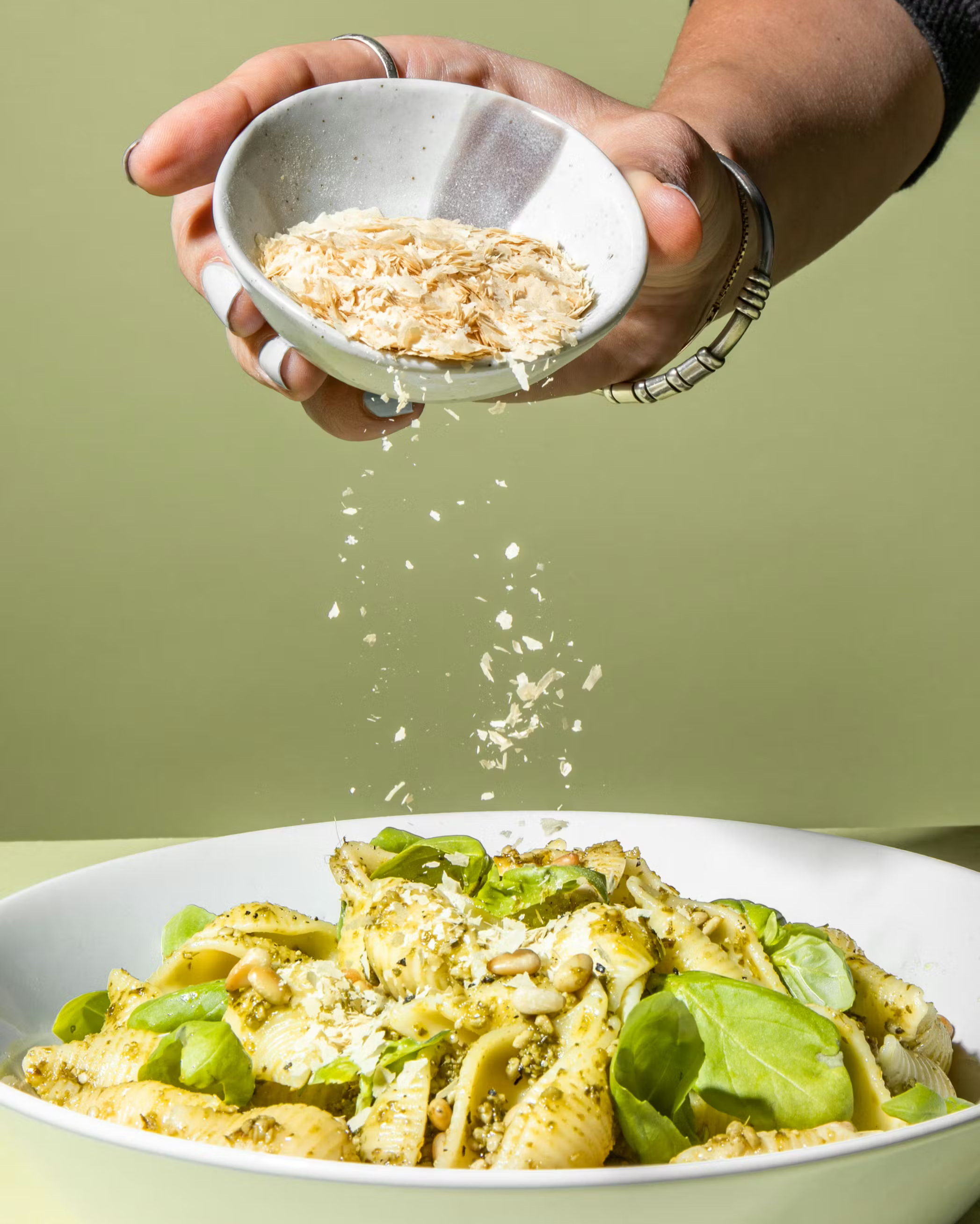 Eine Hand streut Hefeflocken aus einer kleinen Schale über einen Teller Pasta mit Pesto, frischem Basilikum und Pinienkernen.