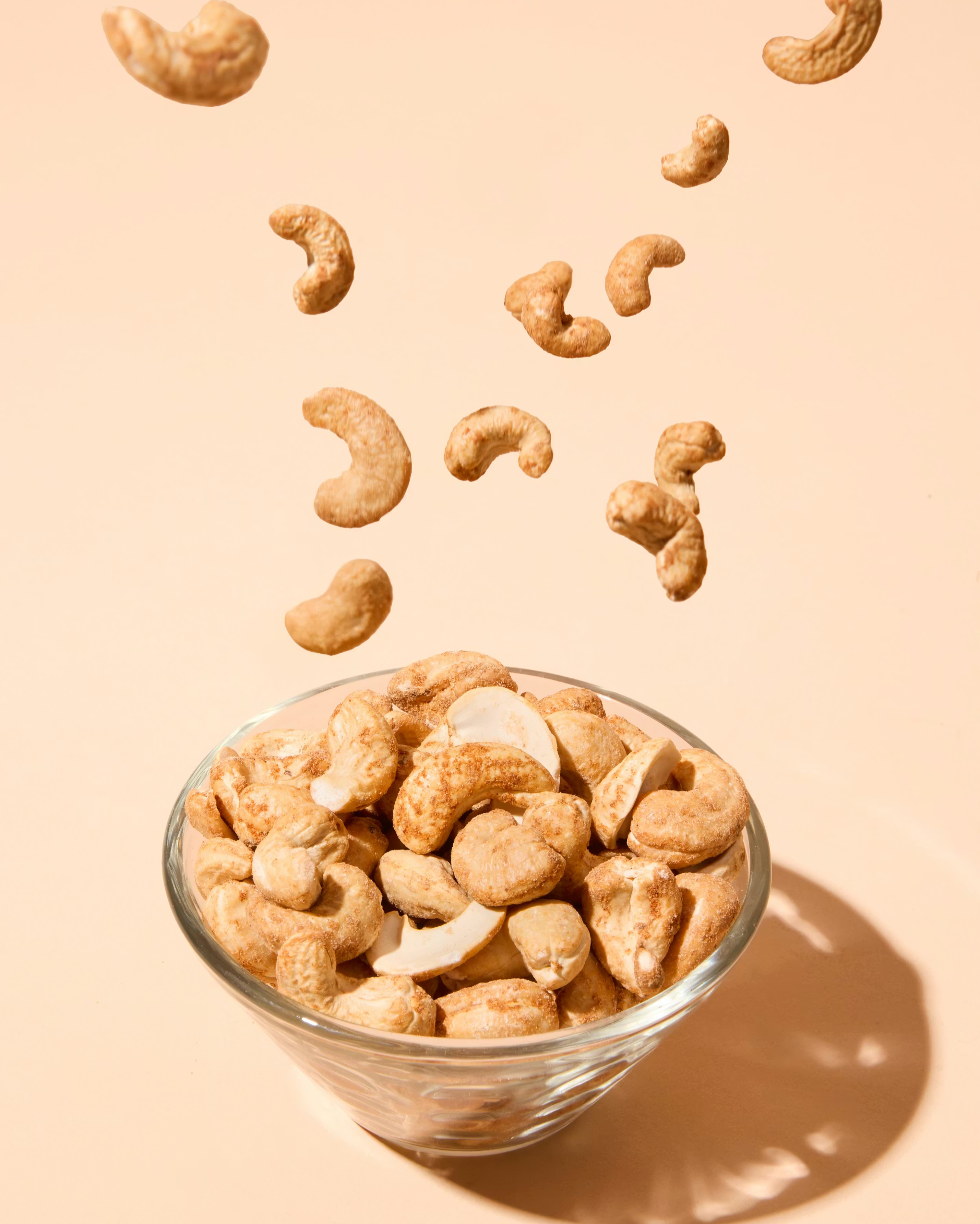Cashewkerne mit Chili-Gewürz fallen in eine Glasschale vor einem beigefarbenen Hintergrund.