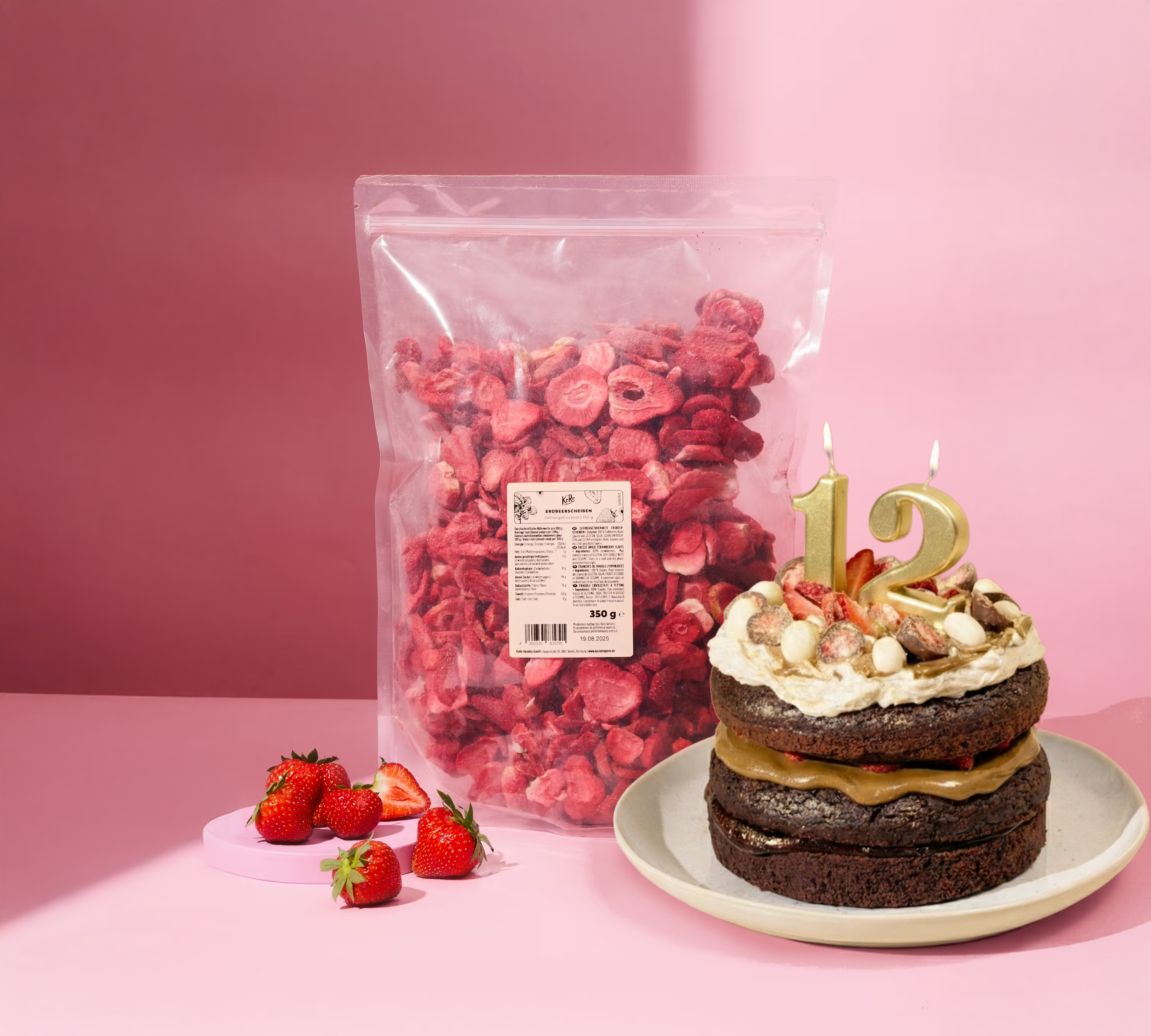 Schokoladige Geburtstagstorte mit goldenen Zahlenkerzen „12“, dekoriert mit frischen Erdbeeren sowie gefriergetrockneten Erdbeeren in Milchschokolade und weißer Schokolade. Daneben steht eine große transparente 350-g-Tüte mit gefriergetrockneten Erdbeerscheiben, dazu frische Erdbeeren vor rosafarbenem Hintergrund.