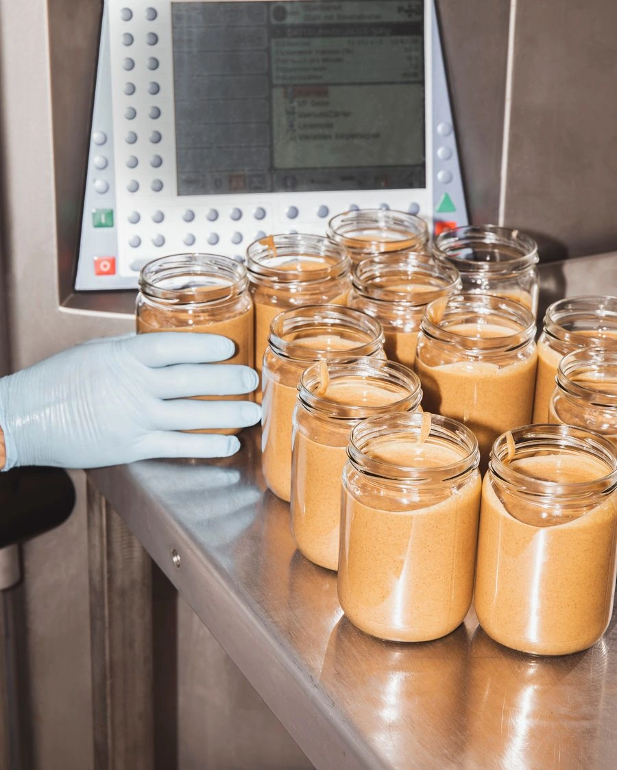 Auf einem Metalltisch in einem sterilen Lagerraum stehen Nussmusgläser – mit Inhalt, aber ohne Etikett. Eine Hand, die einen Einweghandschuh trägt, greift nach einem der Gläser.