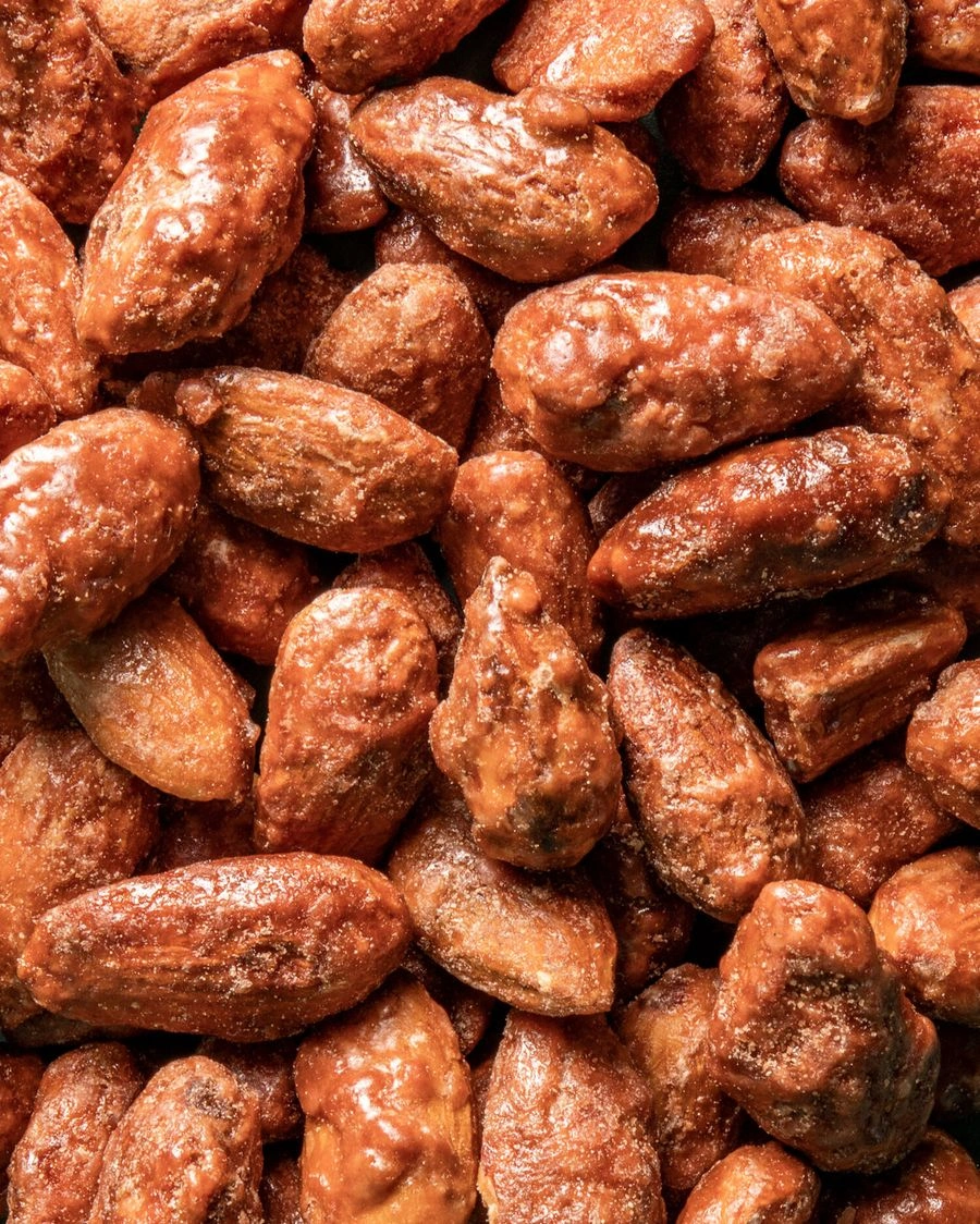 Ein detailliertes Nahaufnahmebild von einem Haufen gebrannter Mandeln. Sie sind alle mit einer glänzenden, karamellisierten Zuckerschicht überzogen und haben eine leicht unebene, knusprige Textur.