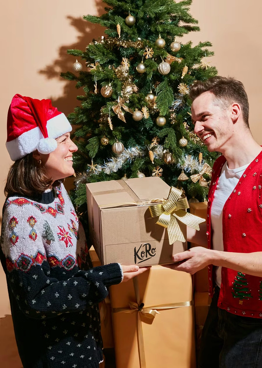 Ein lachendes Paar in weihnachtlicher Kleidung, das gemeinsam ein großes, in braunem Karton verpacktes Geschenk mit dem "KoRo"-Logo und einer goldenen Schleife hält. Die Frau trägt eine Nikolausmütze und einen Norwegerpullover, der Mann einen roten Strick-Pullunder mit Weihnachtsmotiven. Hinter ihnen steht ein geschmückter Weihnachtsbaum und eine Stapel weiterer, in Goldpapier verpackter Geschenke.