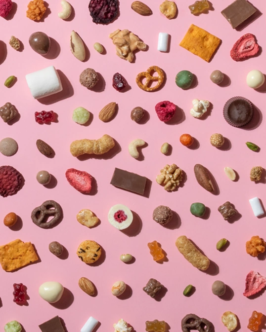  Eine vielfältige Auswahl an Snacks, darunter Nüsse, Gummibärchen, Schokolade, Brezeln und getrocknete Früchte, ist in einem gleichmäßigen Raster auf einem hellrosa Hintergrund angeordnet. 