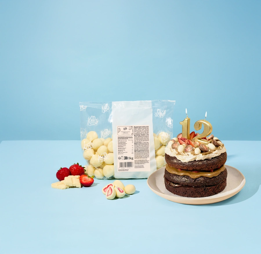 Vegane schokoladige Geburtstagstorte mit goldenen Zahlenkerzen „12“, dekoriert mit gefriergetrockneten Erdbeeren in weißer Schokolade sowie frischen Erdbeeren. Daneben steht eine große 1-kg-Tüte mit gefriergetrockneten Erdbeeren in weißer Schokolade, dazu frische Erdbeeren und Stücke weißer Schokolade vor hellblauem Hintergrund.