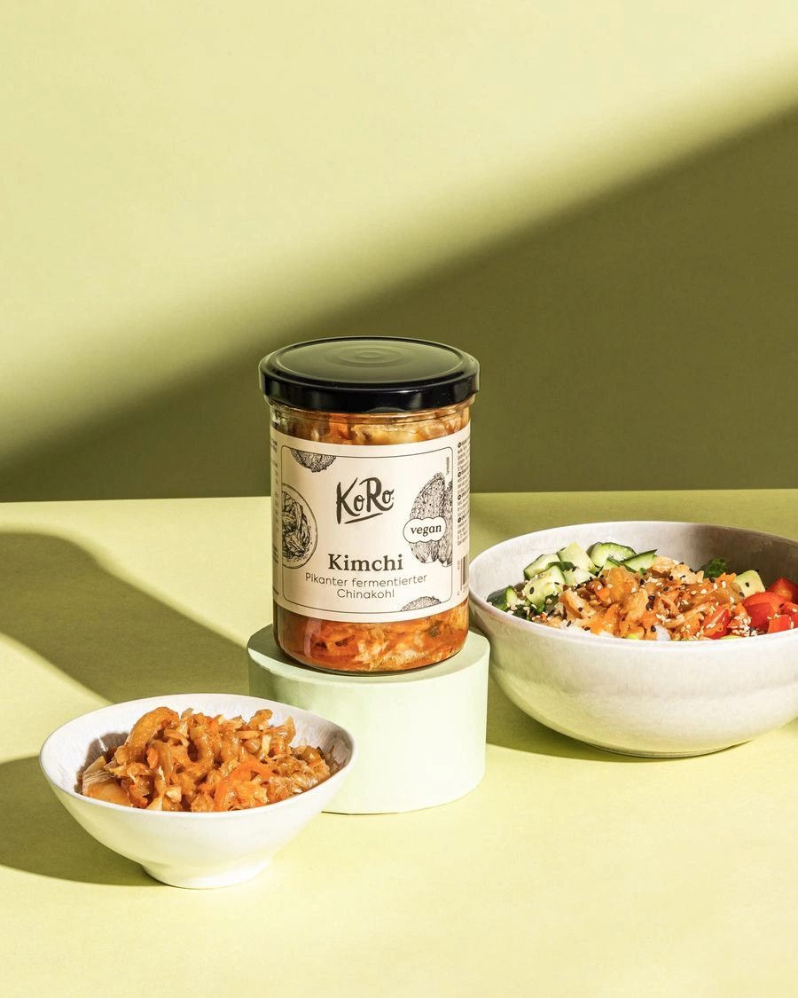 Glas mit Kimchi als Beispiel für fermentierte Lebensmittel, daneben eine Schale mit fermentiertem Kohl und eine Bowl mit frischem Gemüse auf hellgrünem Hintergrund.