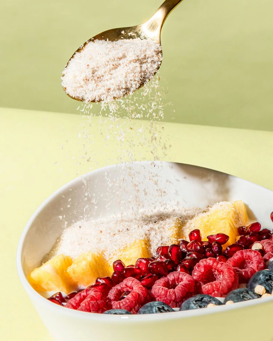 Ein goldener Löffel streut Flohsamenschalen über eine Frühstücksschale mit Ananas, Himbeeren, Heidelbeeren und Granatapfelkernen.