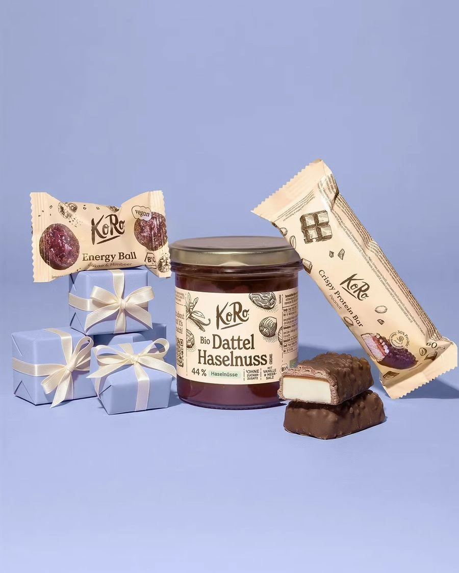 Produktset von KoRo mit einem Glas Bio Dattel-Haselnuss-Creme, einem Energy Ball Kakao & Himbeer und einem Crispy Protein Bar Peanut vor lila Hintergrund mit kleinen Geschenkpäckchen.