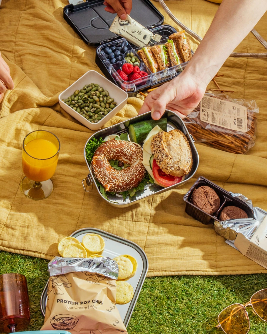 Draufsicht auf ein gemütliches Picknick im Grünen mit belegten Edelstahl-Brotdosen, einem Sesam-Bagel, Sandwiches, frischen Beeren, Edamame und verschiedenen Snacks auf einer gelben Decke.