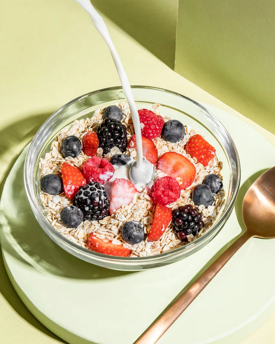 Nahaufnahme: Pflanzliche Milch wird über Bio-Haferflocken Kleinblatt und frische Beeren in einer Glas-Schale gegossen, daneben liegt ein kupferfarbener Löffel.