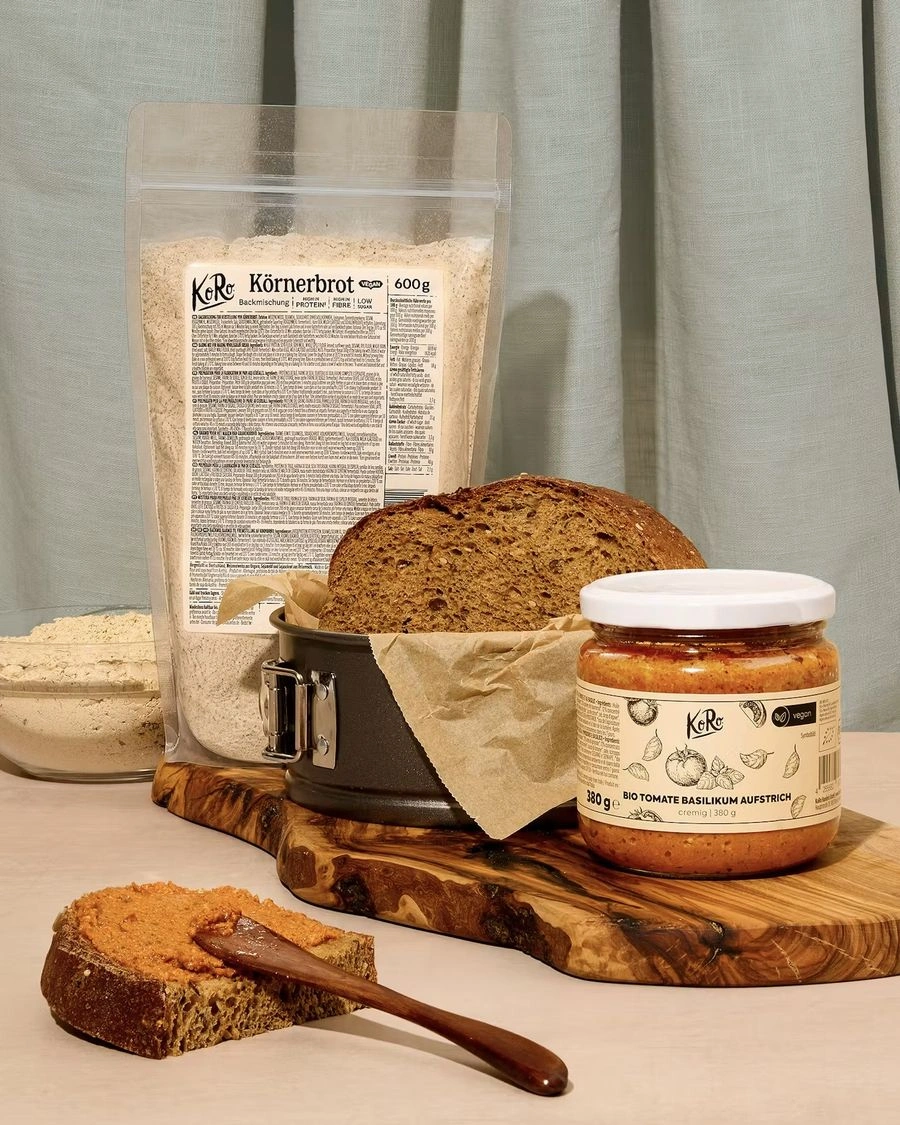 KoRo Körnerbrot Backmischung mit frisch gebackenem Körnerbrot und Bio Tomate-Basilikum-Aufstrich auf einem Holzbrett