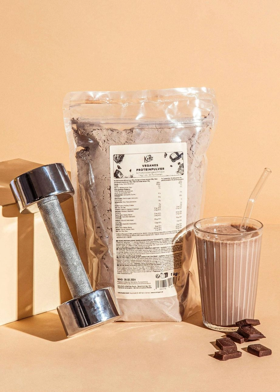 Veganes Schoko-Proteinpulver in einer Tüte vor hellem Hintergrund, davor ist eine Metallhantel an einen Block angelehnt. Auf der rechten Seite steht ein Glas mit einem fertigen Proteinshake und Schokoladenstücke davor verteilt