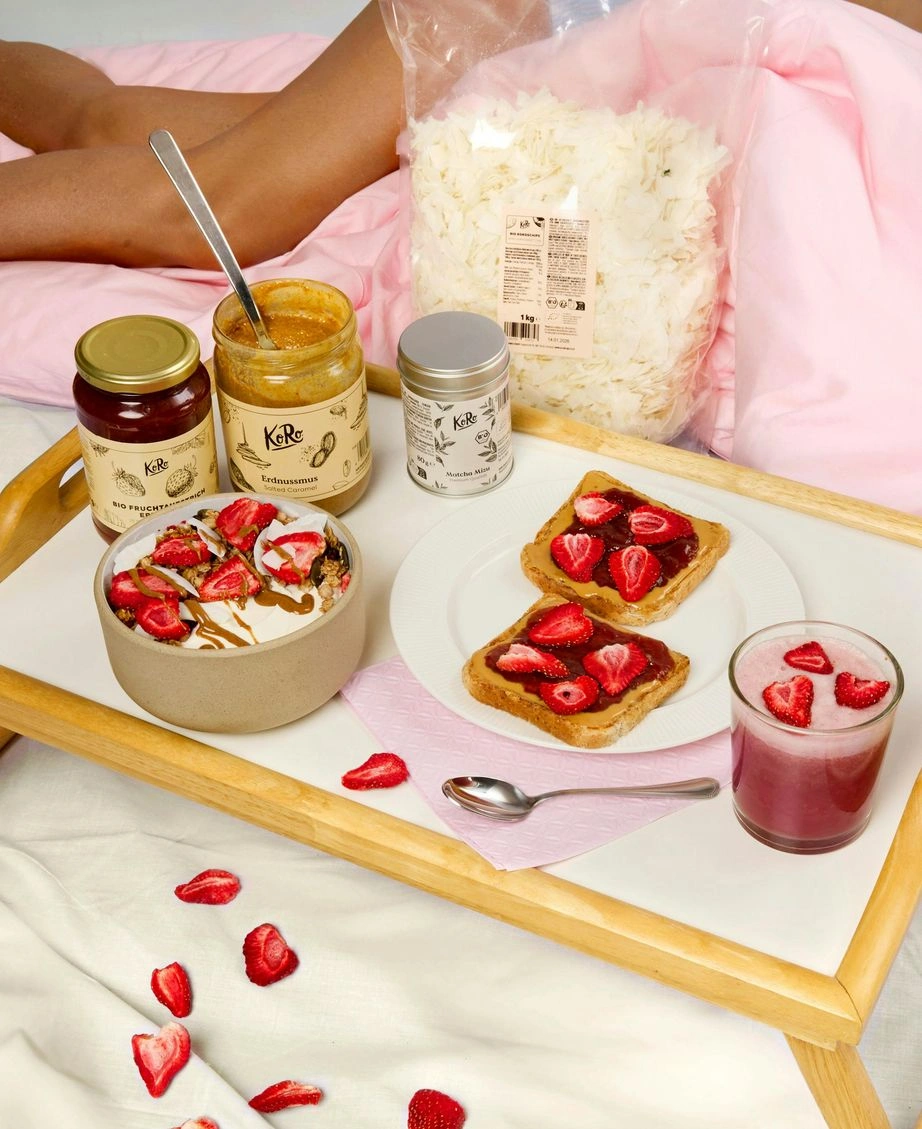 Ästhetisches Frühstück im Bett auf einem hellen Holztablett. Zu sehen sind zwei Scheiben Toast, bestrichen mit Haselnussmus und belegt mit gefriergetrockneten Erdbeerscheiben, eine Bowl mit Joghurt, Granola und Erdbeeren sowie ein Glas rosa Smoothie. Dahinter sind drei Gläser/Dosen der Marke KoRo (Erdnussmus Salted Caramel, Bio-Fruchtaufstrich Erdbeere, Matcha-Mix) und ein 1kg-Beutel Kokoschips arrangiert. Der Hintergrund ist hell und gemütlich mit rosa Bettwäsche.