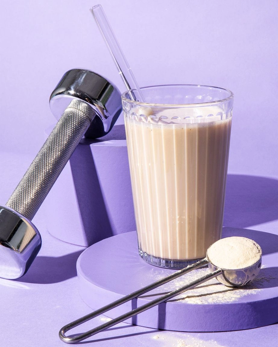 Ein Glas mit cremigem Vanille-Proteinshake und Glasstrohhalm steht auf einem lilafarbenen Podest, daneben ein Messlöffel mit veganem Vanille-Proteinpulver und eine silberne Hantel.