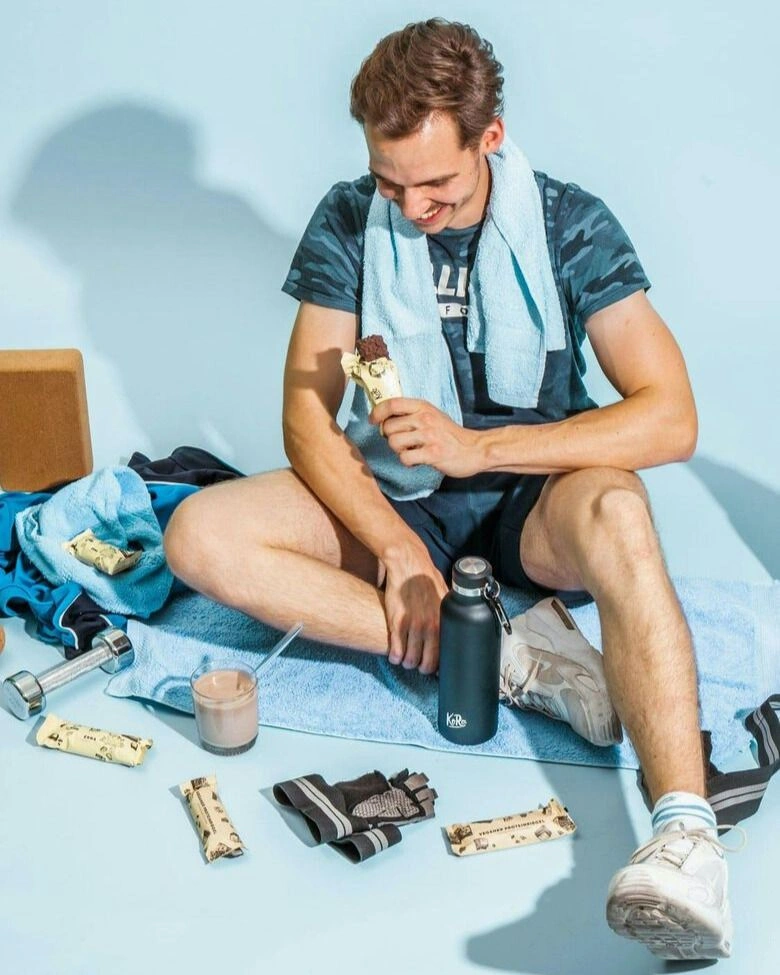 Ein Mann sitzt vor einem hellblauen Hintergrund auf einem Handtuch auf dem Boden. Er hat einen Proteinriegel in der Hand und vor ihm liegen diverse Proteinprodukte sowie Hanteln und eine Wasserflasche. Er selbst trägt Sportkleidung.