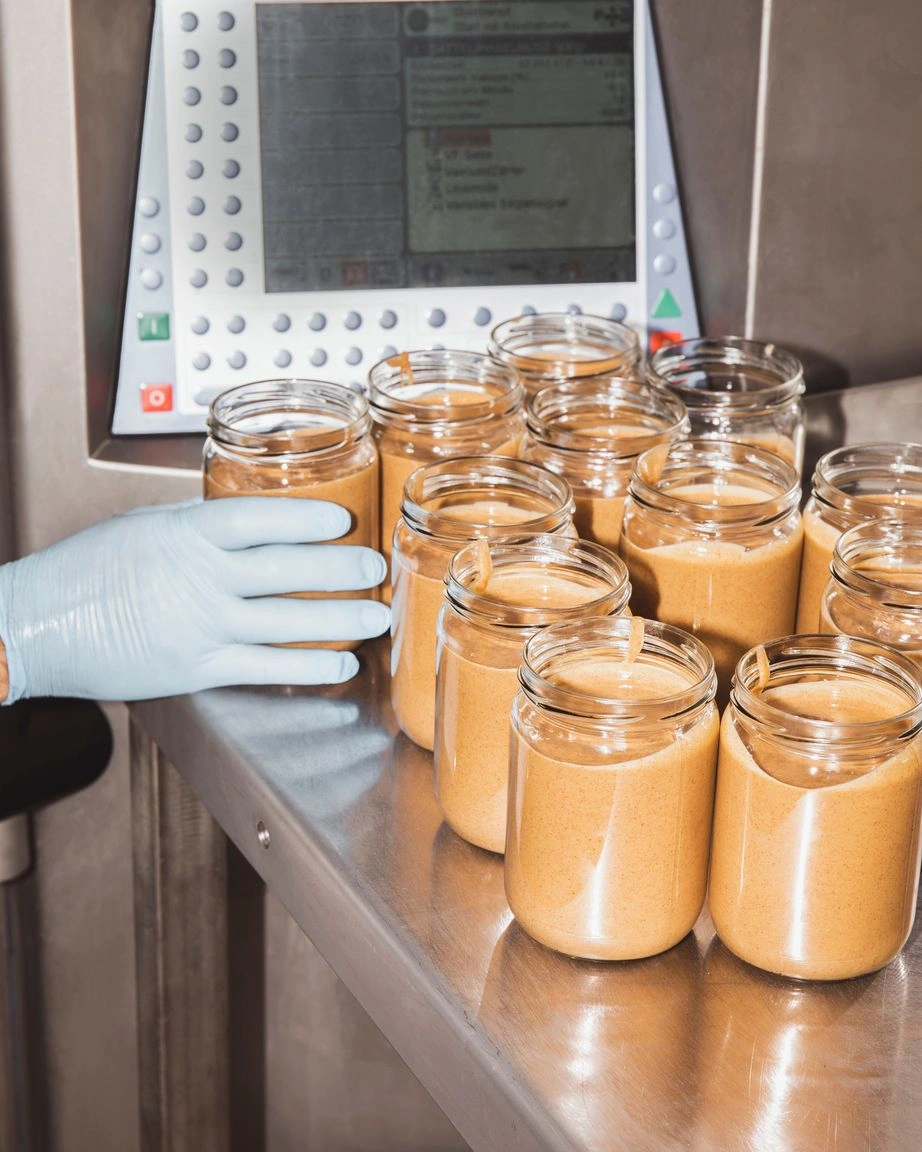 Auf einem Metalltisch in einem sterilen Lagerraum stehen Nussmusgläser – mit Inhalt, aber ohne Etikett. Eine Hand, die einen Einweghandschuh trägt, greift nach einem der Gläser.