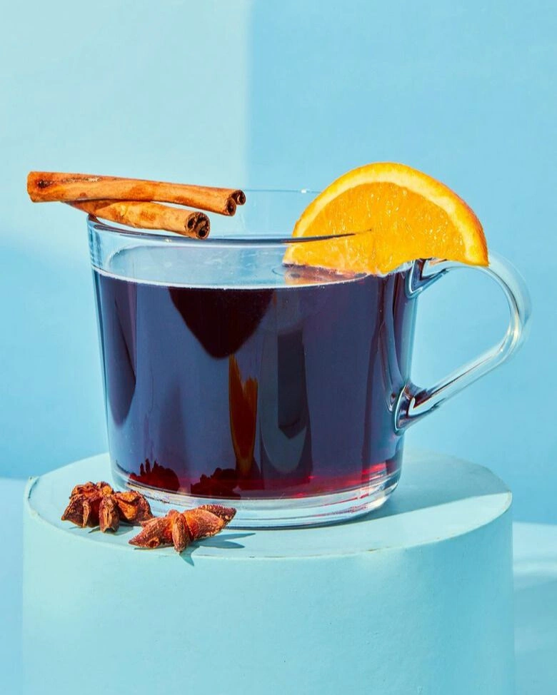 Ein Glas Glühwein oder Punsch, garniert mit einer Orangenscheibe und Zimtstangen, steht auf einem hellblauen Podest. Sternanis liegt daneben.