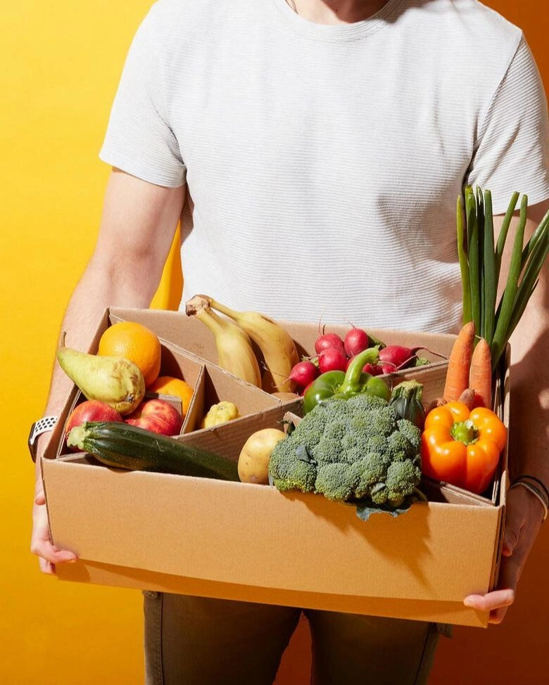  Ein Mann in einem weißen T-Shirt hält eine braune Pappkiste mit frischem Obst und Gemüse in der Hand. Die Kiste ist in mehrere Fächer unterteilt und enthält unter anderem Brokkoli, Bananen, Karotten, Paprika, Zucchini, Äpfel und Birnen. Der Mann steht vor einem leuchtend gelben Hintergrund.